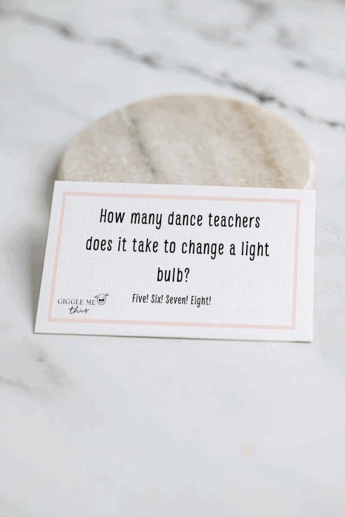 A card on a marble surface reads: “How many dance teachers does it take to change a light bulb? Five! Six! Seven! Eight!”—one of the classic jokes about dance.