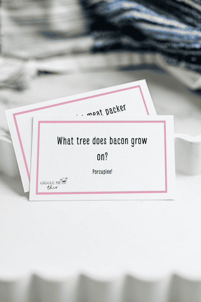 A white card with a pink border reads, What tree does bacon grow on? Porcupine! This playful Bacon Jokes card sits in a white tray, with another card partially visible and a blue-striped cloth in the background.