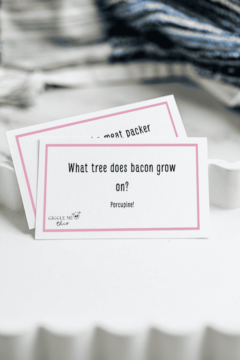 A white card with a pink border reads, What tree does bacon grow on? Porcupine! This playful Bacon Jokes card sits in a white tray, with another card partially visible and a blue-striped cloth in the background.